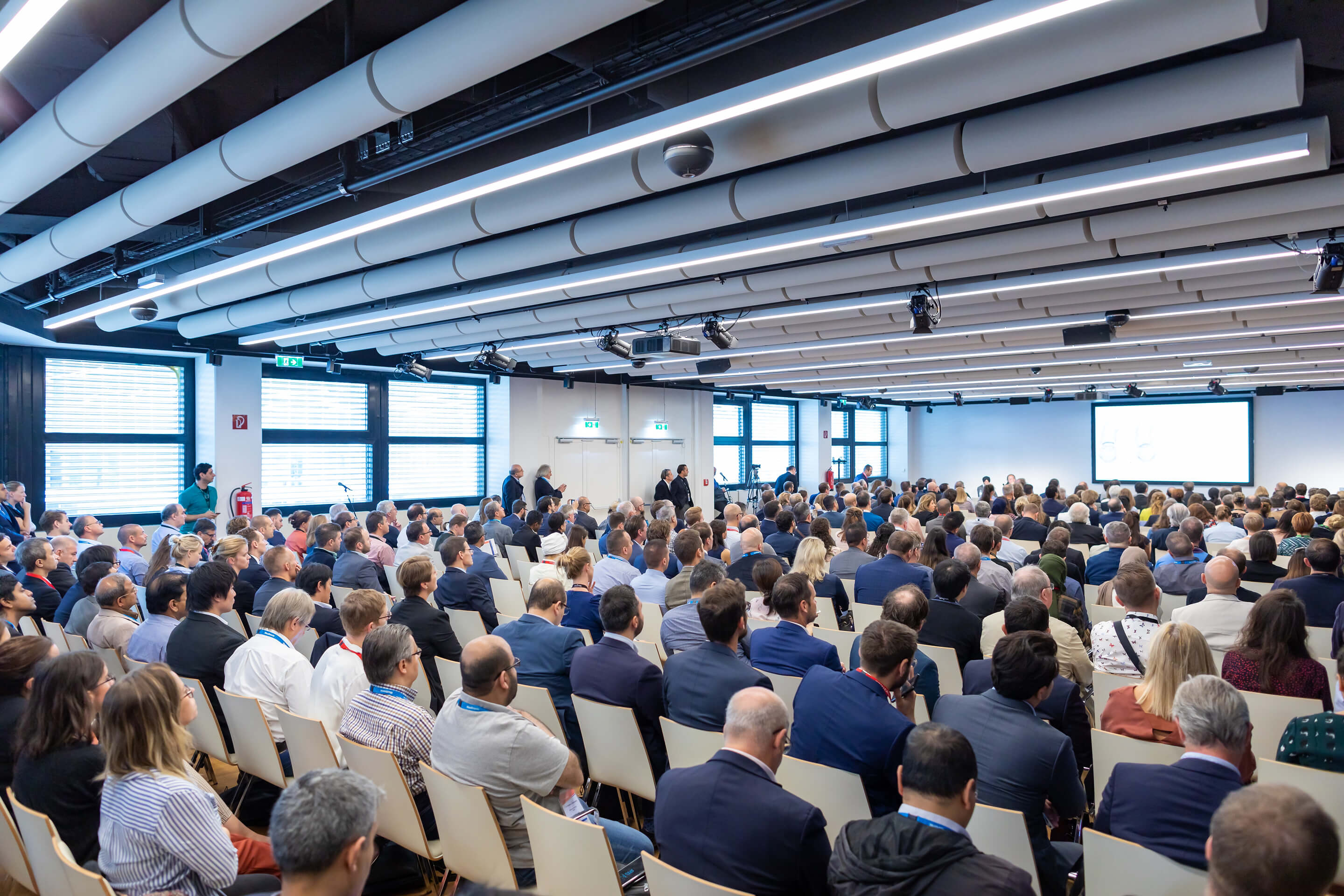Large audience seated in a conference room facing a presentation screen at the front.