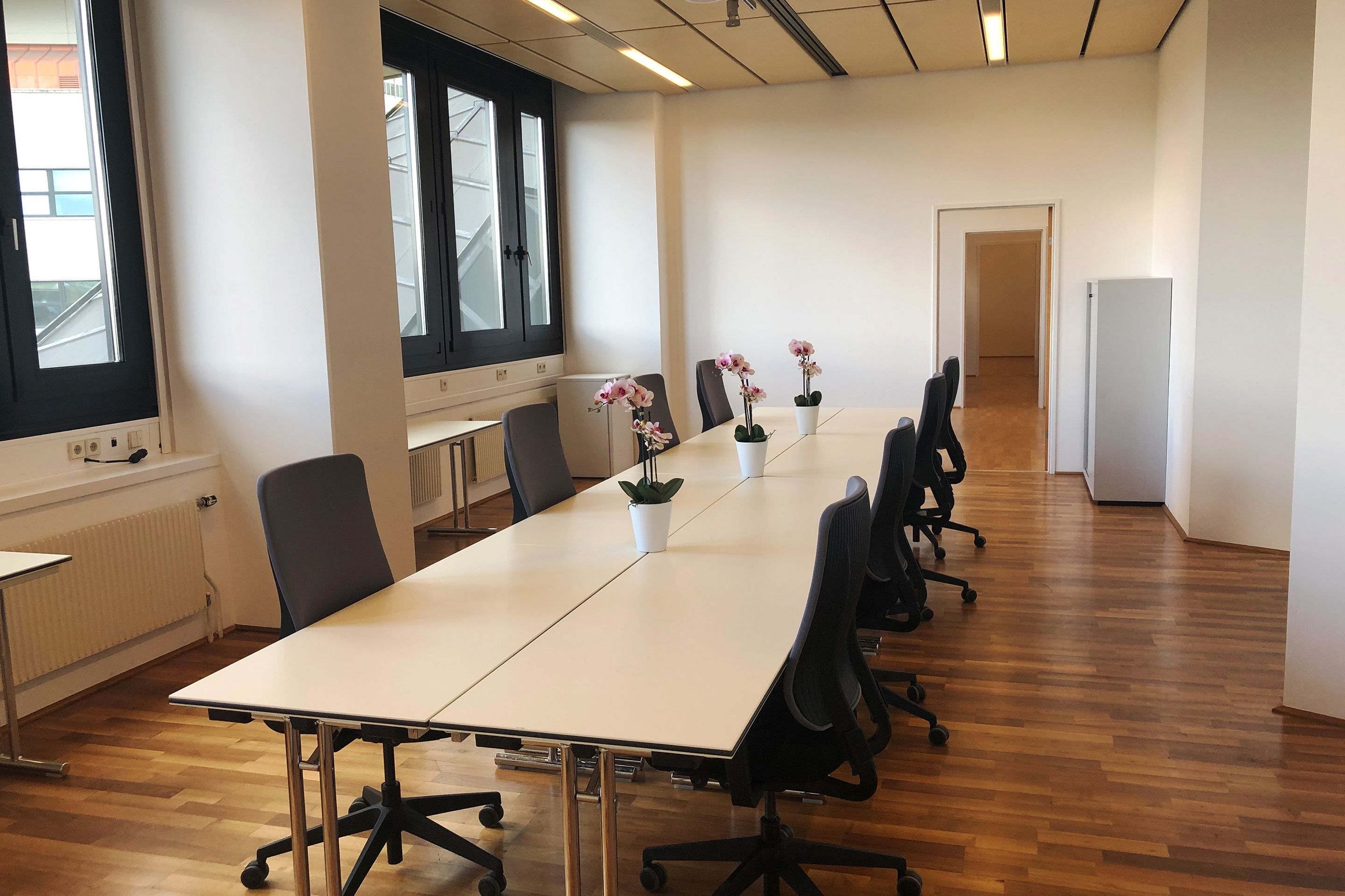 Konferenzraum mit langem Tisch, acht Bürostühlen und vier Orchideen in weißen Töpfen