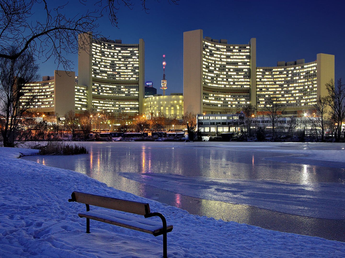 Bank am schneebedeckten Ufer eines zugefrorenen Sees vor beleuchteten Hochhäusern bei Nacht