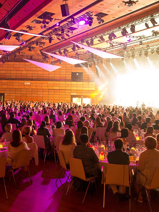 Large audience seated at tables in a brightly lit event hall with stage lighting