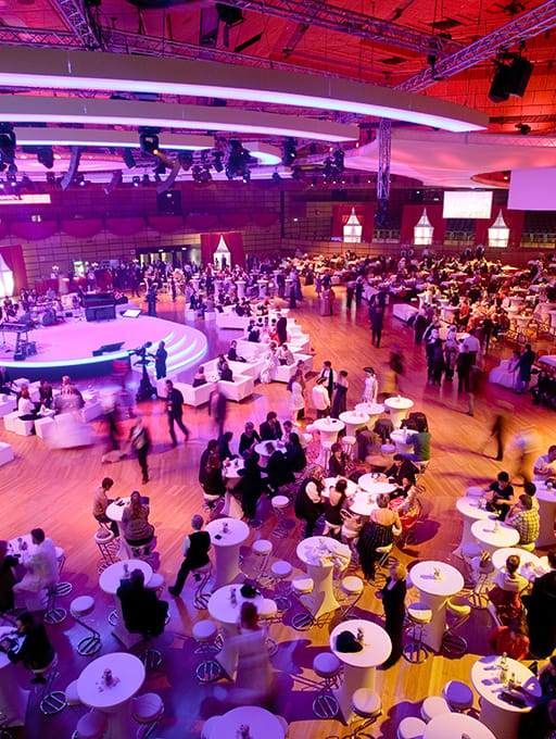 Large indoor event with many people seated at round tables and a stage with lighting above