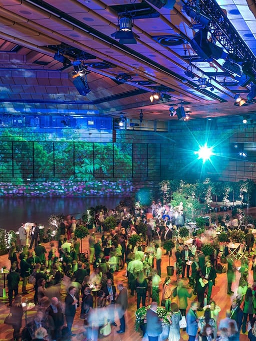 Large indoor event with many people and potted plants under blue and green lighting