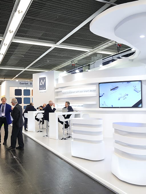 Messestand mit weißen Möbeln, mehreren Personen im Gespräch und Bildschirm an der Wand