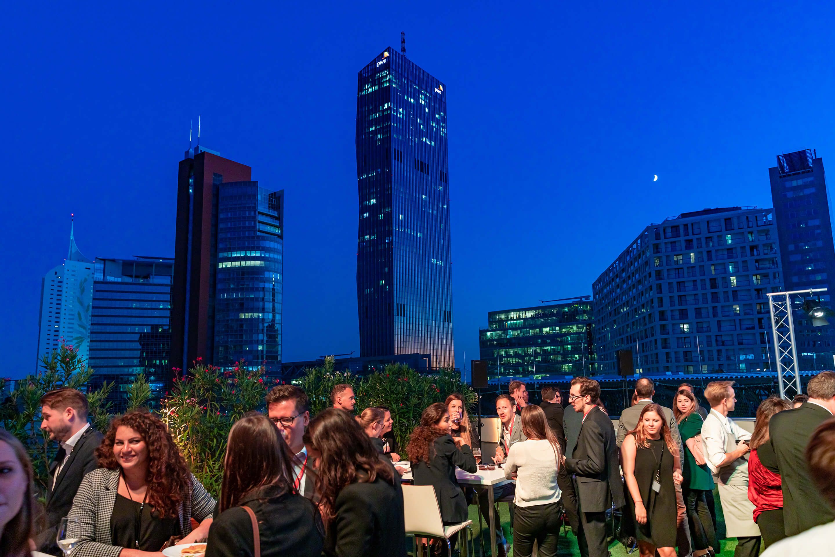 Menschen bei Abendveranstaltung auf Dachterrasse vor beleuchteter Stadtsilhouette