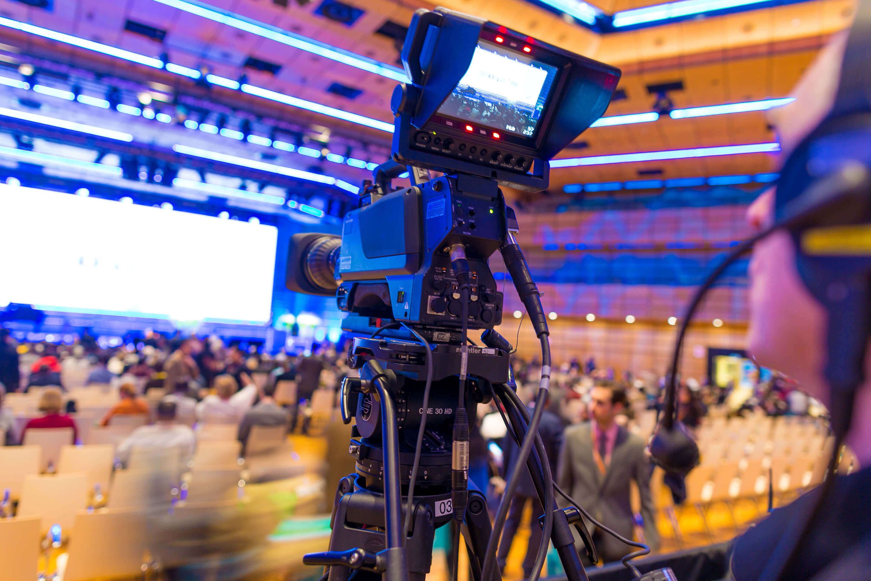 Professional video camera recording an event in a large auditorium with seated audience