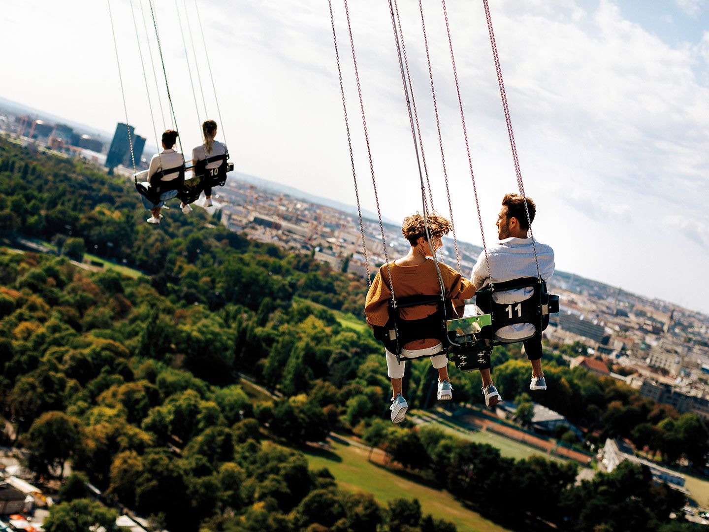 Four people sitting on numbered swing seats high above a city and green park below