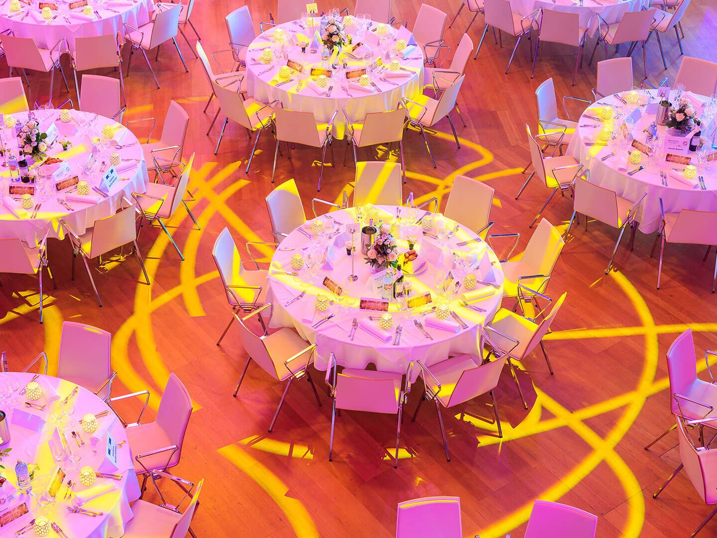 Round tables with white tablecloths and chairs set for an event with floral centerpieces and yellow lighting patterns on the floor