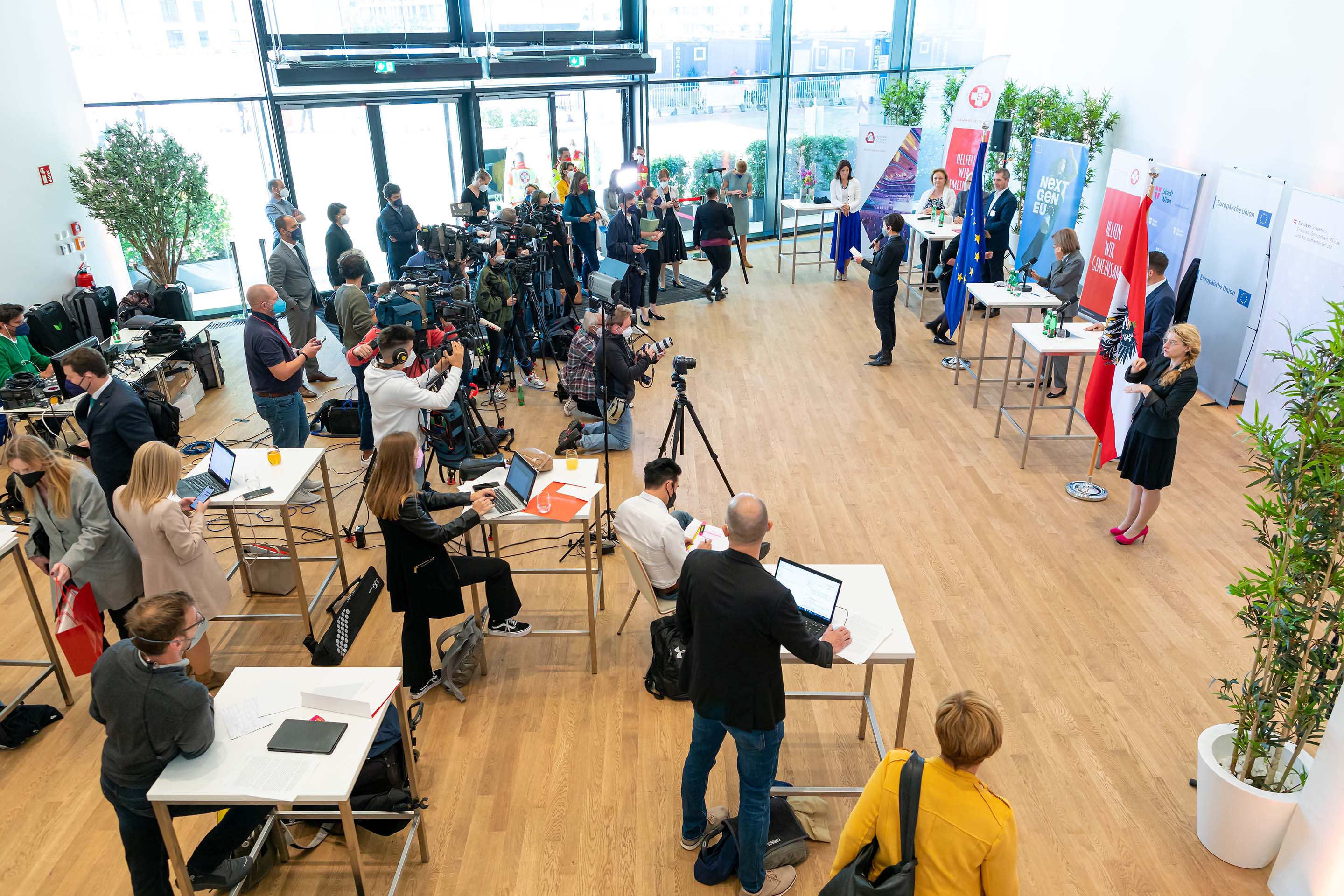 Pressekonferenz mit mehreren Personen, Kameras, EU- und Österreich-Flaggen und Dolmetscherin in Gebärdensprache