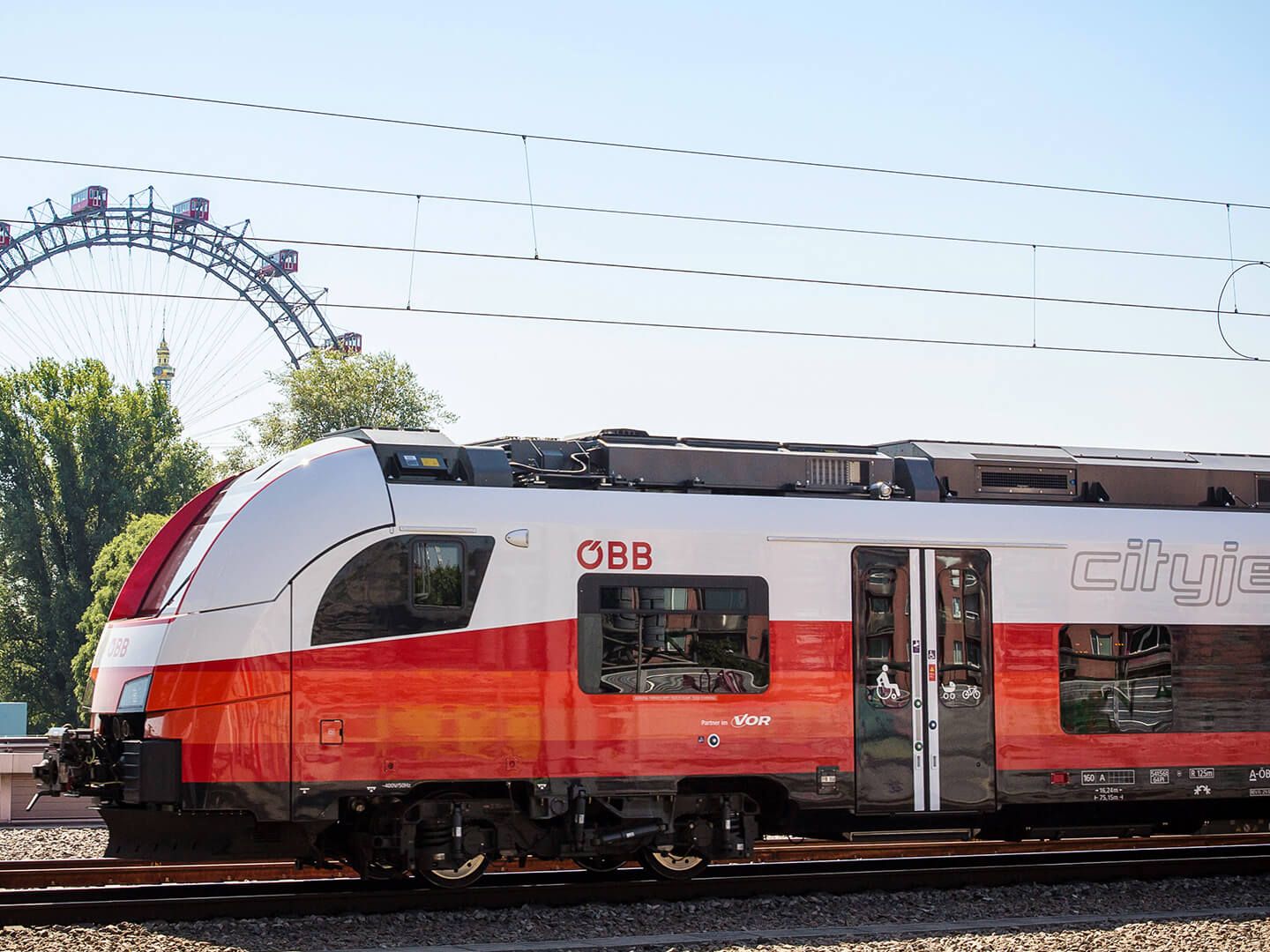 ÖBB-Zug in Rot-Weiß auf Gleisen mit Riesenrad und Bäumen im Hintergrund