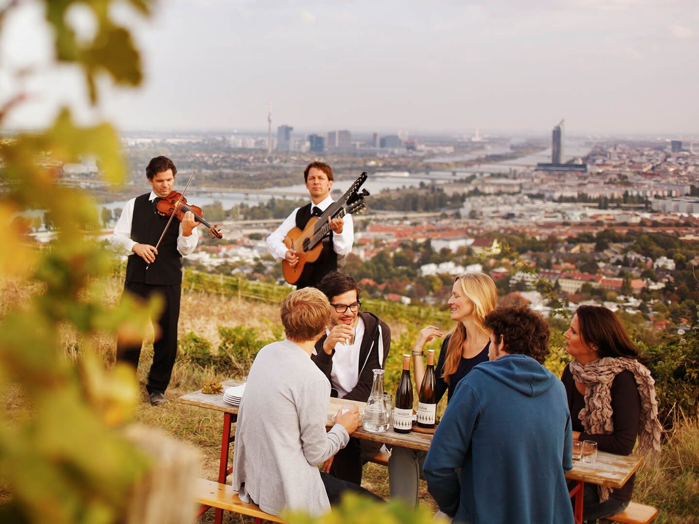 Menschen sitzen an einem Tisch im Freien mit Weinflaschen, Musiker spielen Geige und Gitarre.