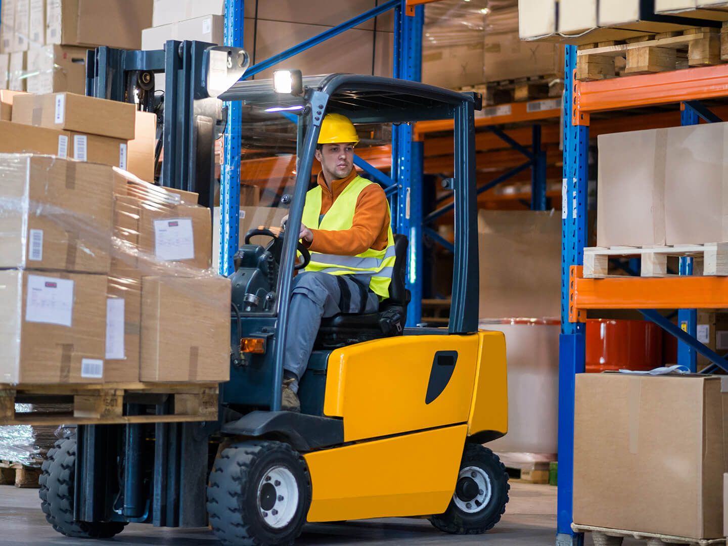 Worker in safety vest and helmet operating yellow forklift with stacked boxes in warehouse