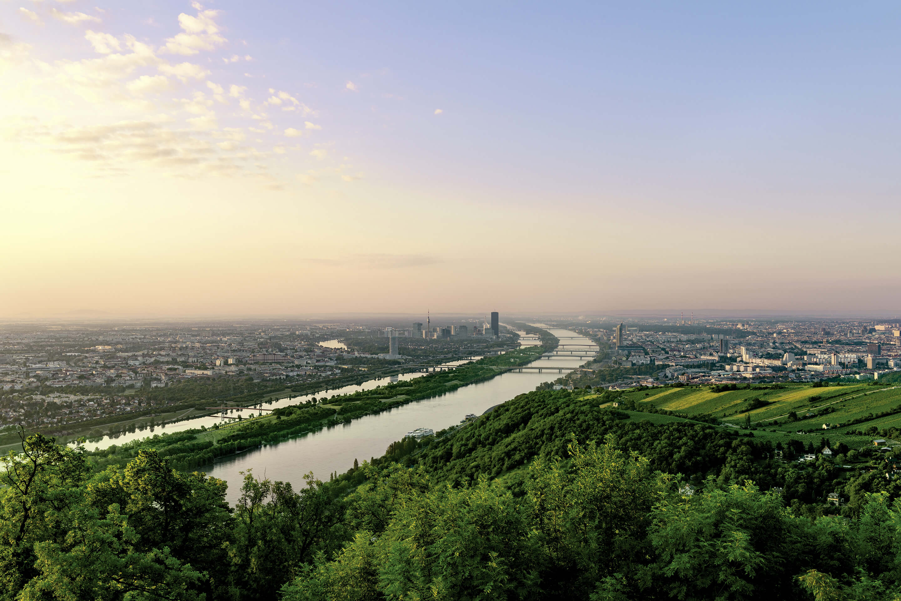 Fluss mit mehreren Brücken, umgeben von Stadt und grünen Hügeln bei Sonnenuntergang.