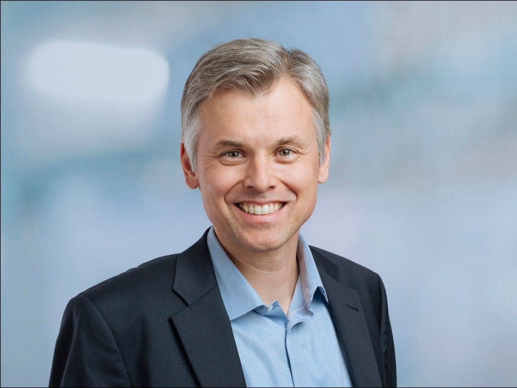 Man wearing a black blazer and light blue collared shirt against a blue background