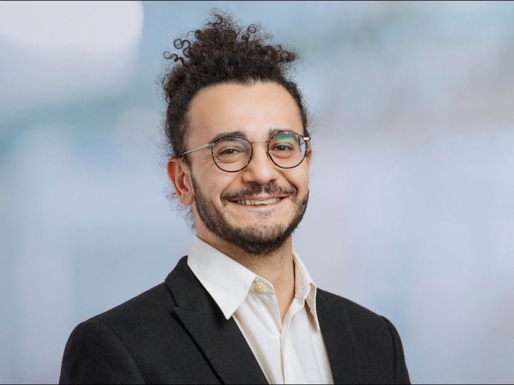 Person with curly hair wearing a black blazer and white collared shirt against a blurred background