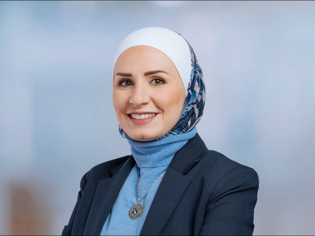 Person wearing a patterned headscarf, blue turtleneck, dark blazer, and round pendant necklace