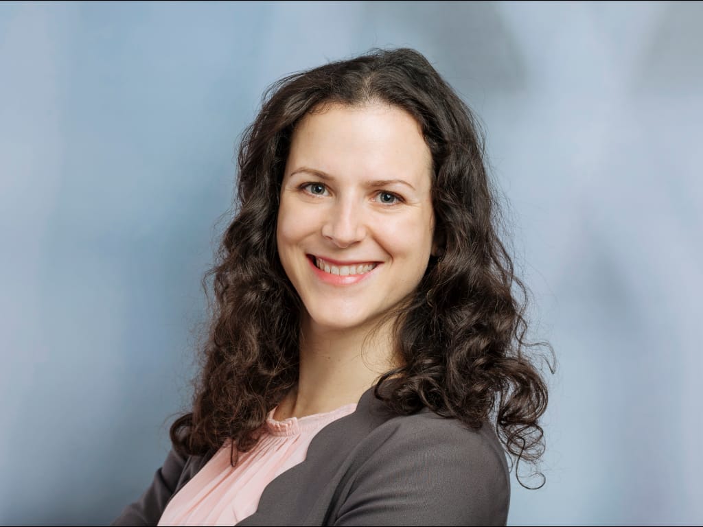 Person with long curly hair wearing a gray blazer and light pink blouse against a blue background