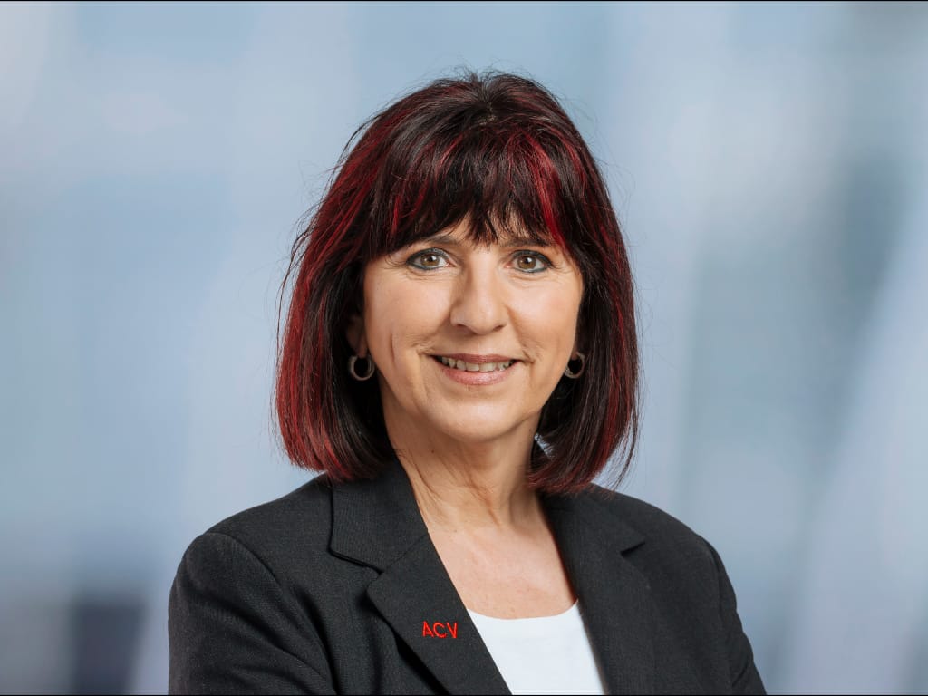 Person with shoulder-length dark hair with red highlights wearing a black blazer with red "ACV" embroidery and a white top against a blurred blue-gray background