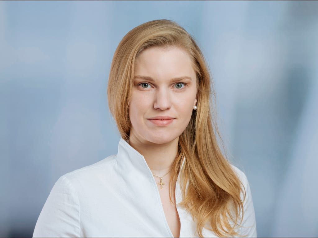 Person with long blonde hair wearing a white shirt and a gold cross necklace against a blue background