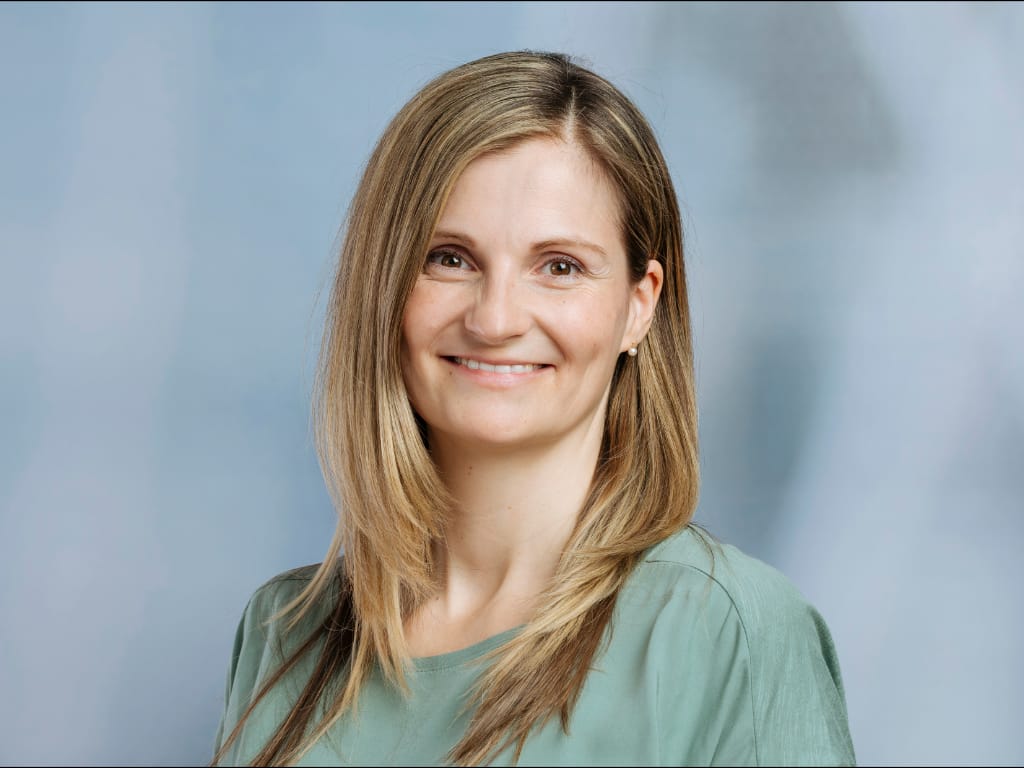 Frau mit langen, glatten blonden Haaren und grünem Oberteil vor blauem Hintergrund