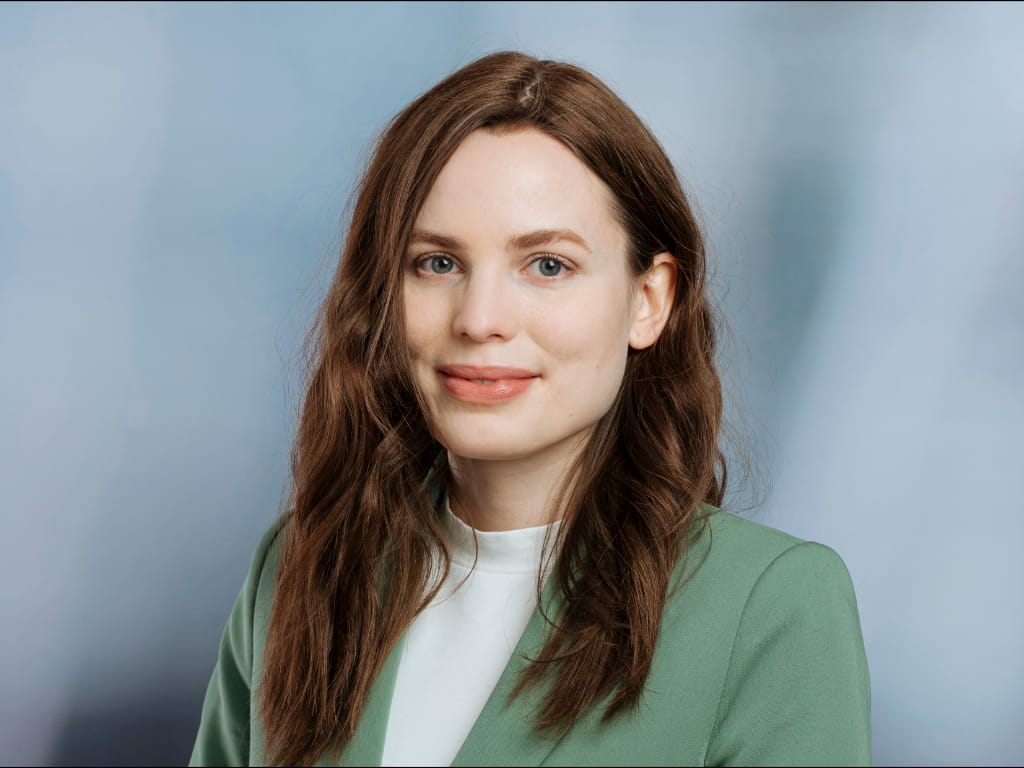 Frau mit langen braunen Haaren trägt grünen Blazer und weißes Oberteil vor blauem Hintergrund