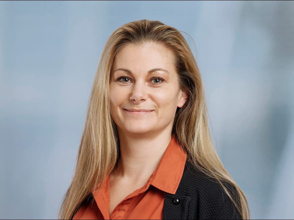 Person with long blonde hair wearing an orange shirt and black cardigan against a blue background