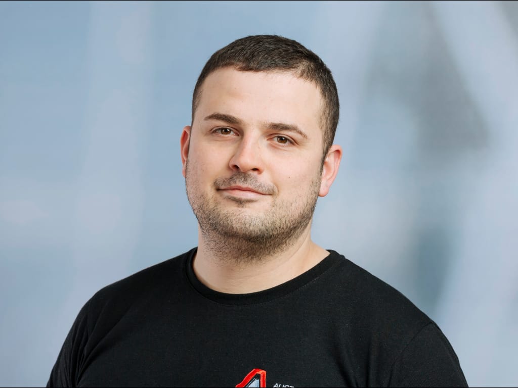 Man with short dark hair and beard wearing a black shirt with a red and white logo on a light blue background