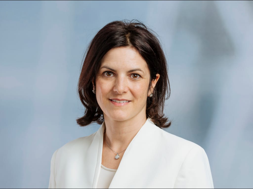 Person with shoulder-length dark hair wearing a white blazer and a pendant necklace against a blue background