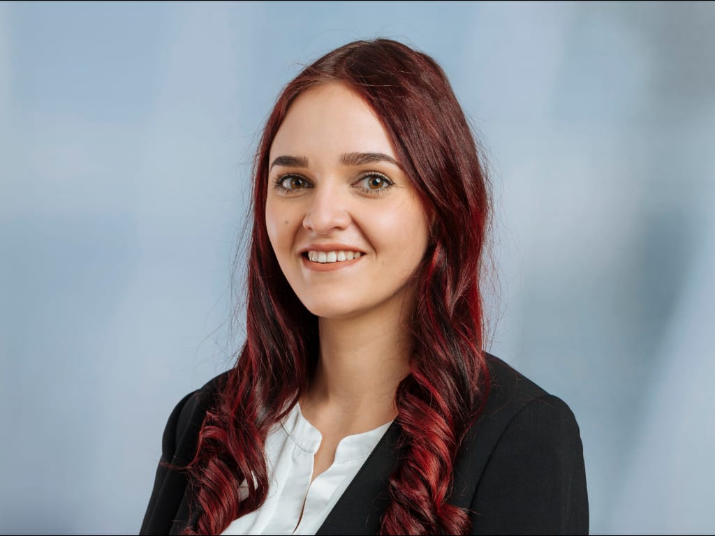 Person with long red curly hair wearing a black blazer and white shirt against a plain background