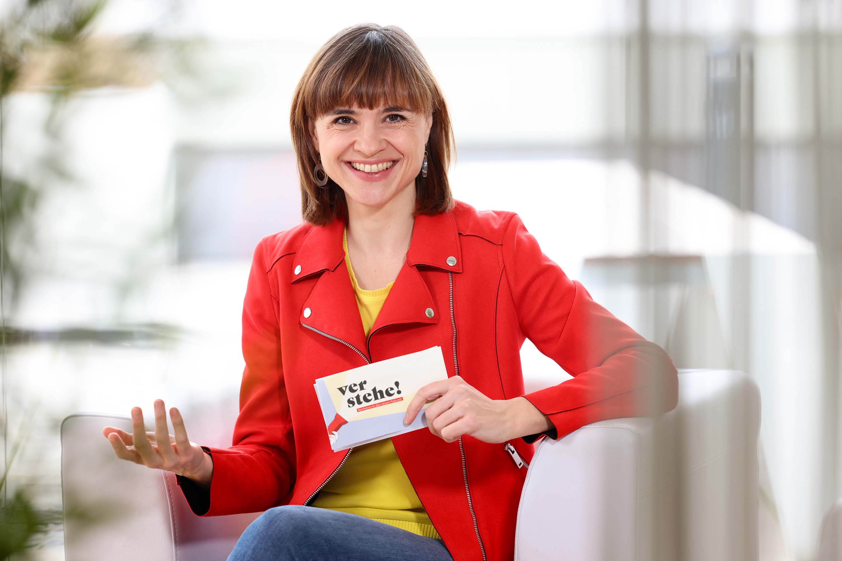 Person in roter Jacke hält Karte mit Text „verstehe!“ in der Hand, sitzt auf weißem Sessel.
