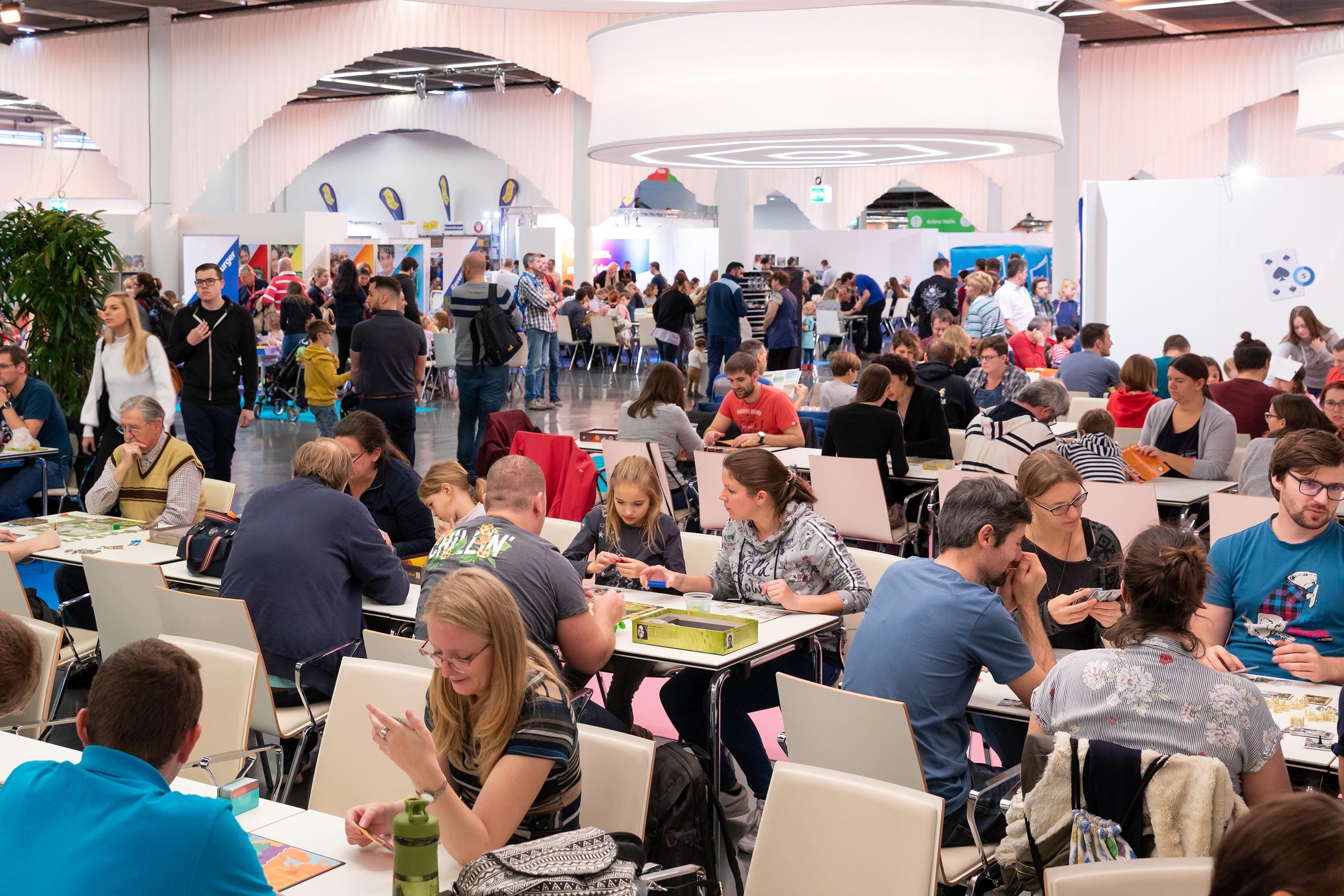 Viele Menschen sitzen an Tischen und spielen Gesellschaftsspiele in einer großen Halle.