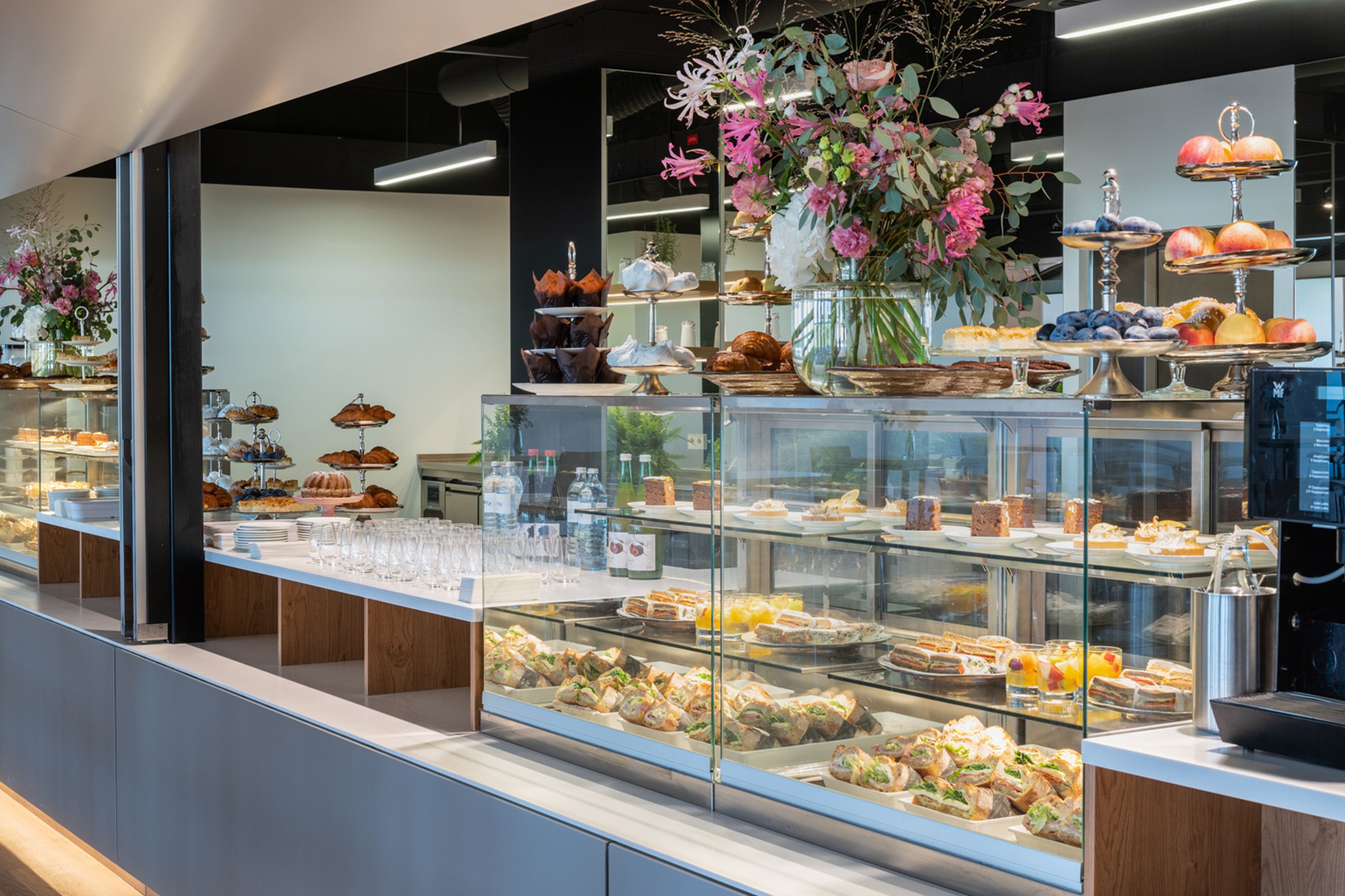 Theke mit Glasvitrinen, gefüllt mit Sandwiches, Kuchen und Obst, Blumenarrangement obenauf