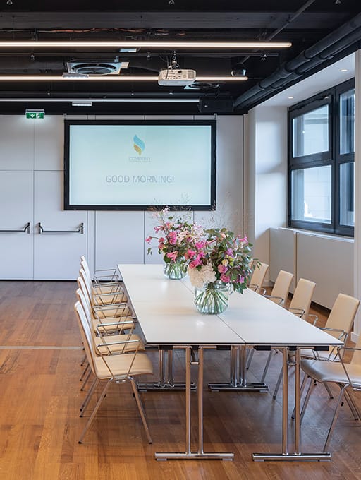 Konferenzraum mit Tisch, Stühlen, Blumenvasen und Leinwand mit Text „GOOD MORNING!“