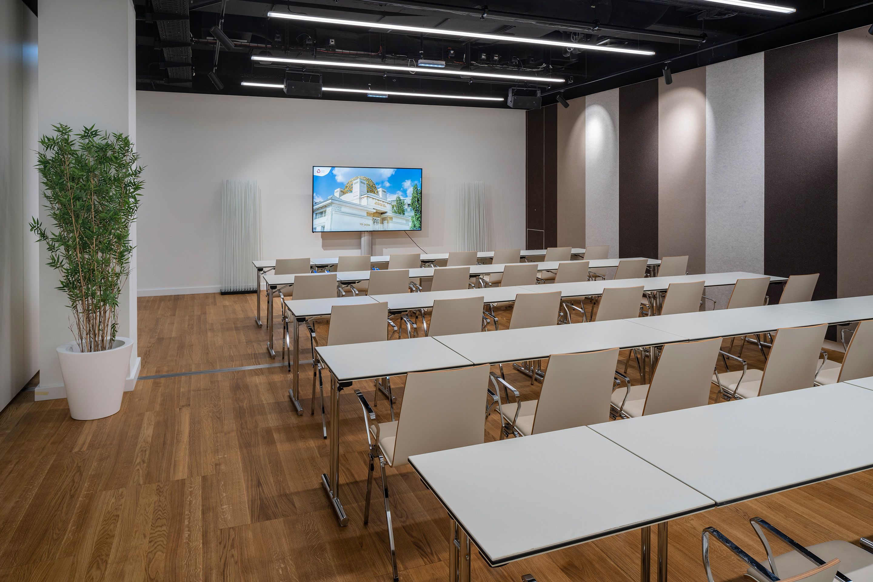 Konferenzraum mit Reihen von Tischen und Stühlen, Bildschirm an der Wand, Pflanze links