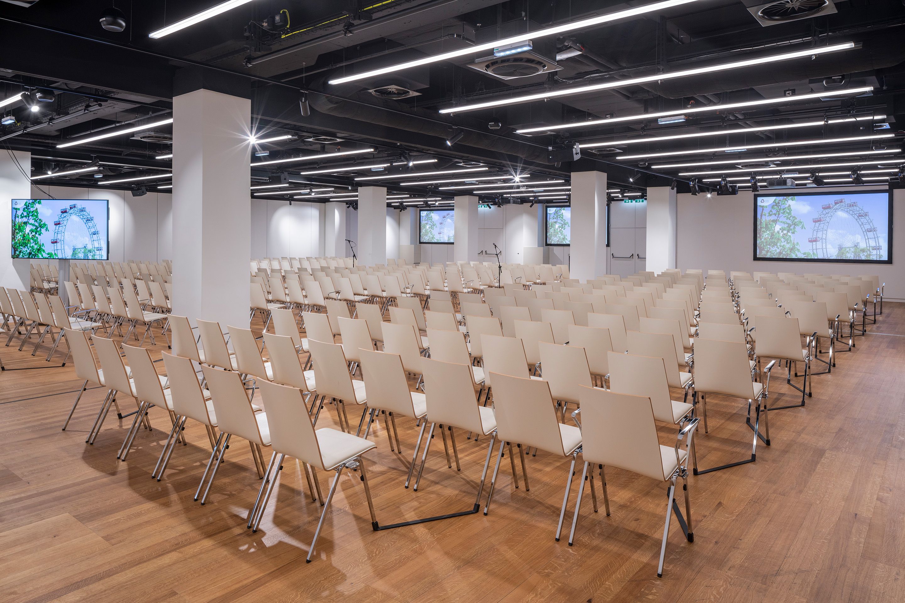 Leerer Konferenzraum mit vielen weißen Stühlen und drei Bildschirmen an der Wand.