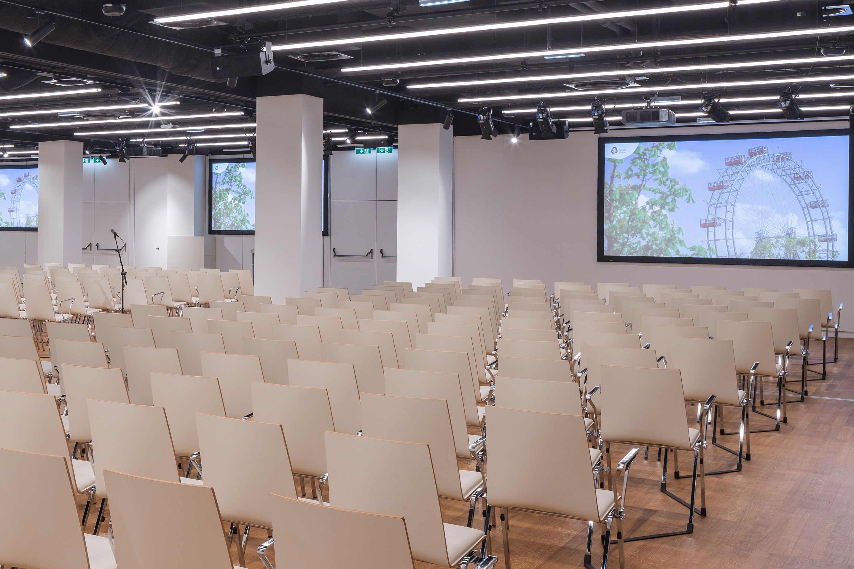 Leerer Konferenzraum mit Reihen von beigen Stühlen und zwei Bildschirmen mit Riesenrad-Motiv.