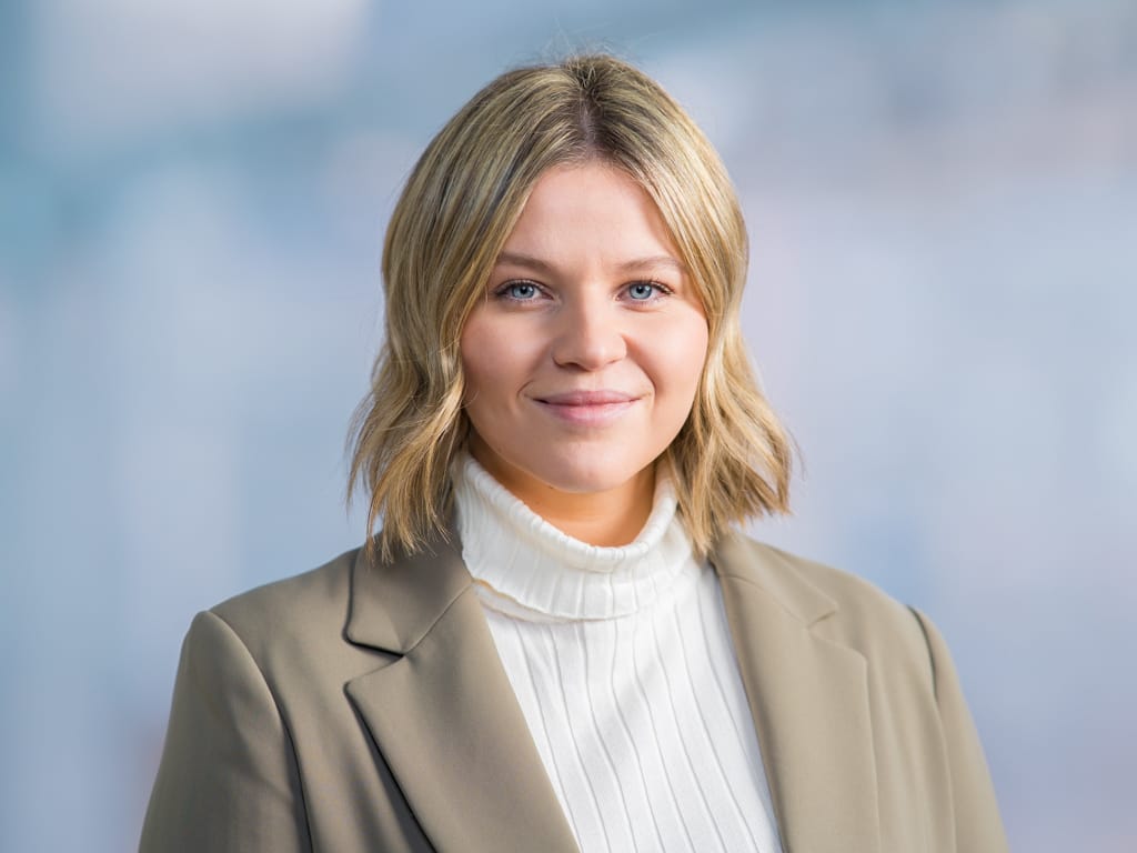 Person with shoulder-length blonde hair wearing a white turtleneck and beige blazer against a blurred background