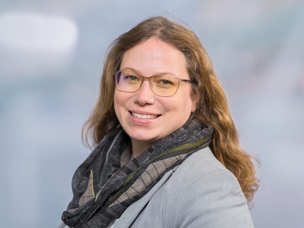 Person with long wavy hair wearing a gray blazer and patterned scarf against a blurred background