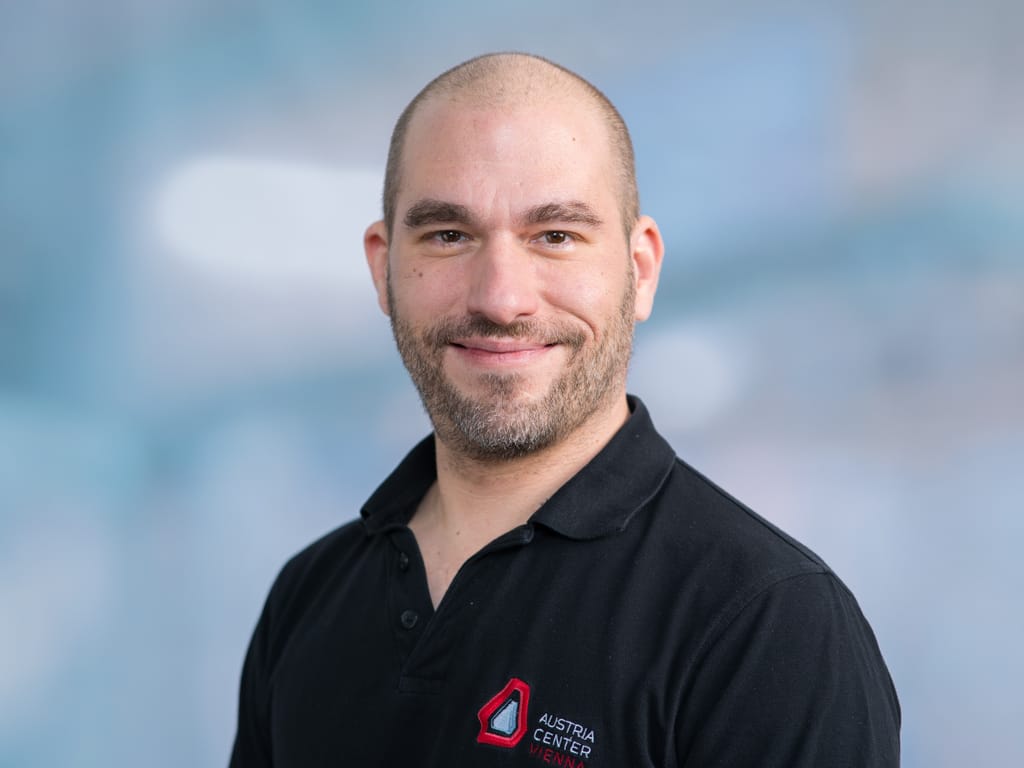 Man wearing black polo shirt with Austria Center Vienna logo on left chest against blurred blue background