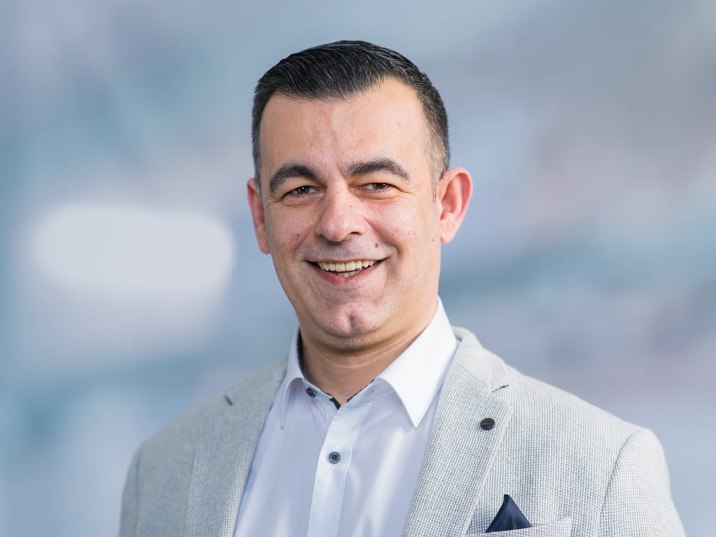 Man wearing a light gray blazer and white shirt with a dark pocket square against a blurred background