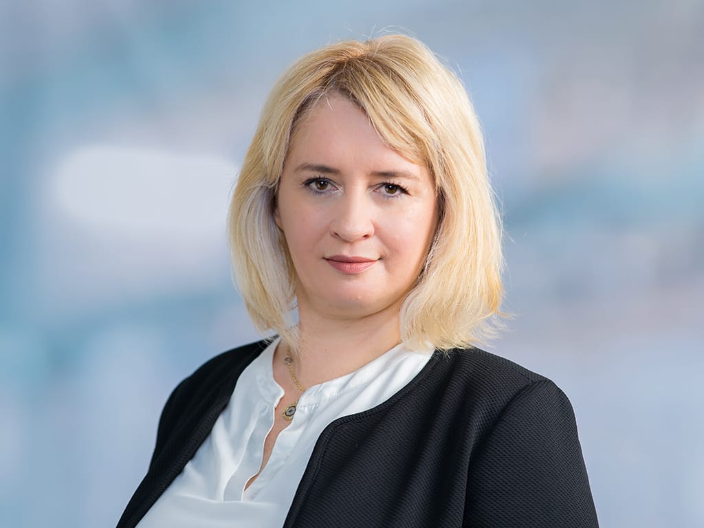 Person with shoulder-length blonde hair wearing a white blouse and black jacket against a blue background