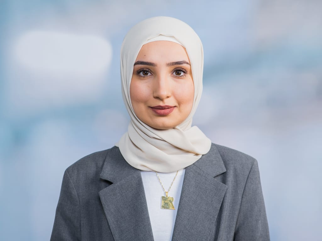 Person wearing a beige hijab, gray blazer, white shirt, and gold pendant necklace