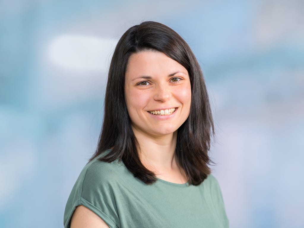 Woman with shoulder-length dark hair wearing a green short-sleeve shirt against a blue background