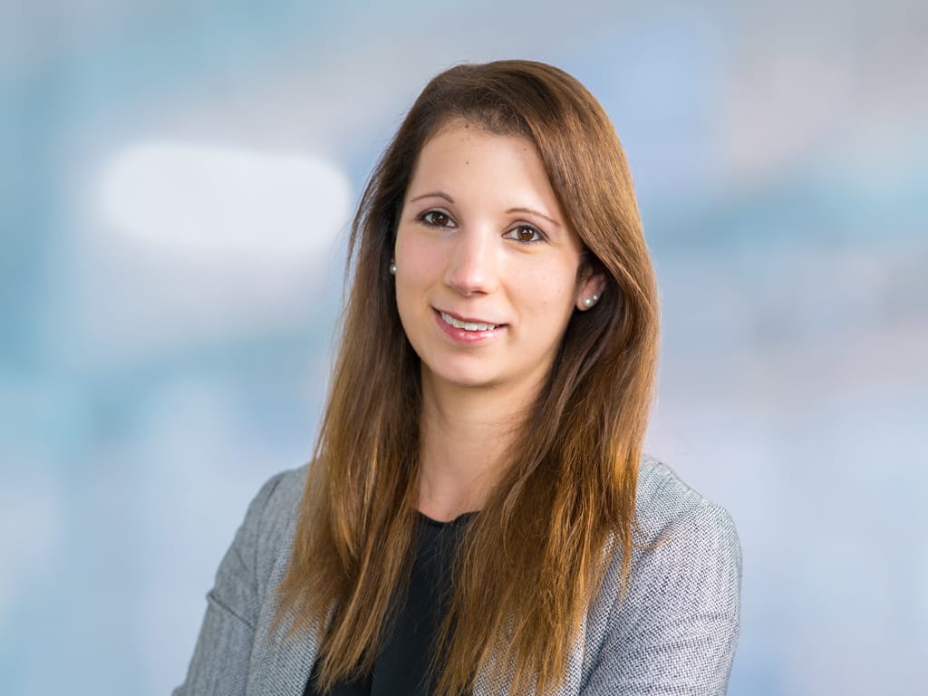 Woman with long brown hair wearing a gray blazer and black top against a blurred background