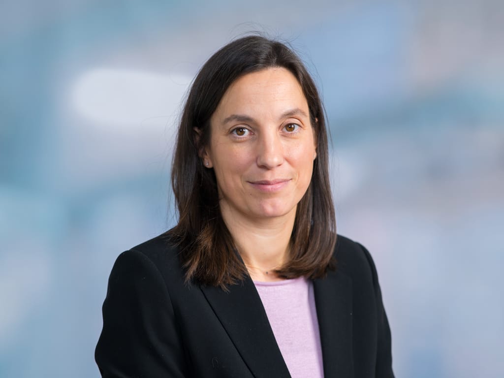 Person with shoulder-length brown hair wearing a black blazer and light purple top against a blue background