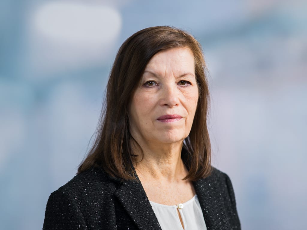 Woman with shoulder-length brown hair wearing a black blazer and white blouse against a blurred background