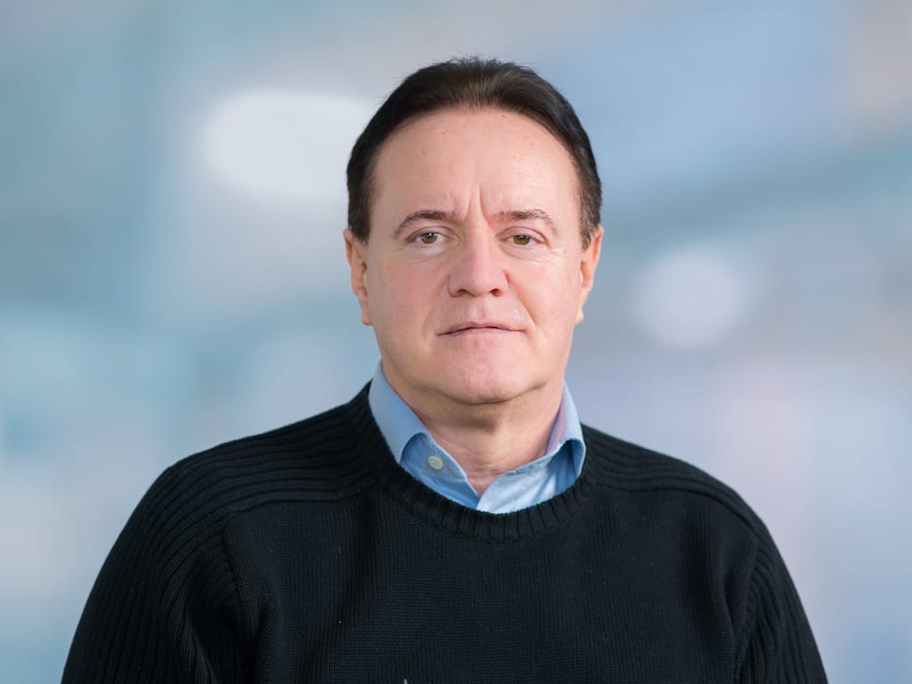 Man wearing a black sweater over a light blue collared shirt against a blurred background