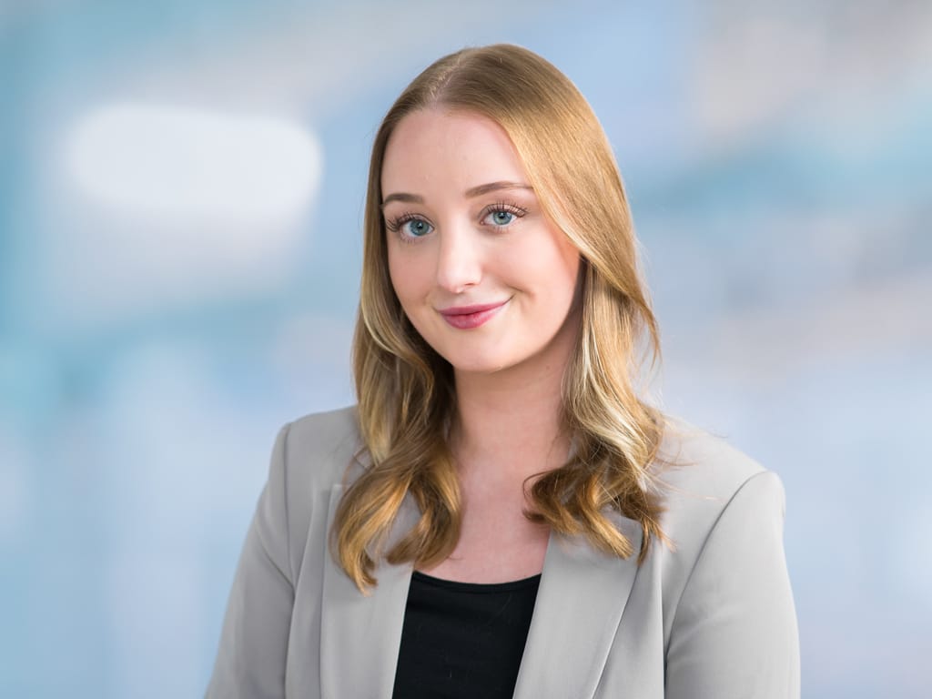 Person with long blonde hair wearing a light gray blazer and black top against a blue background