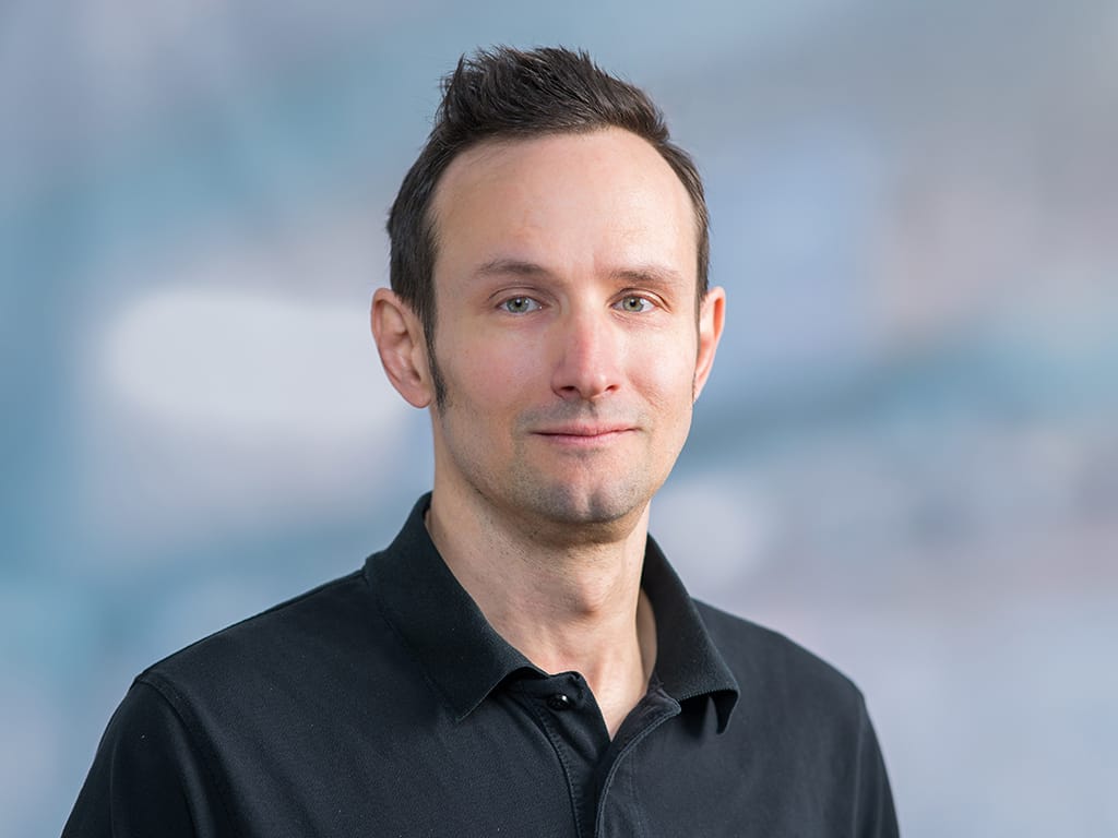 Man wearing a black collared shirt against a blurred blue-gray background