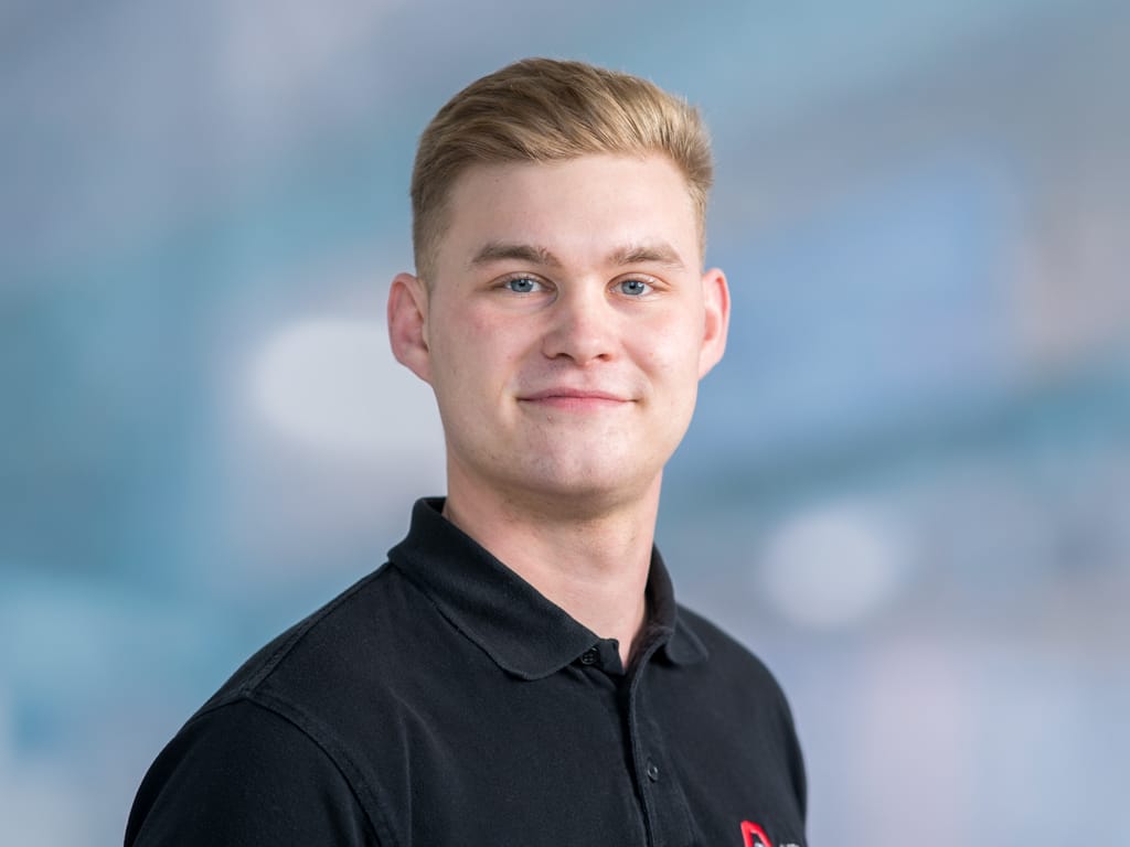 Person with short blond hair wearing a black collared shirt against a blurred blue background