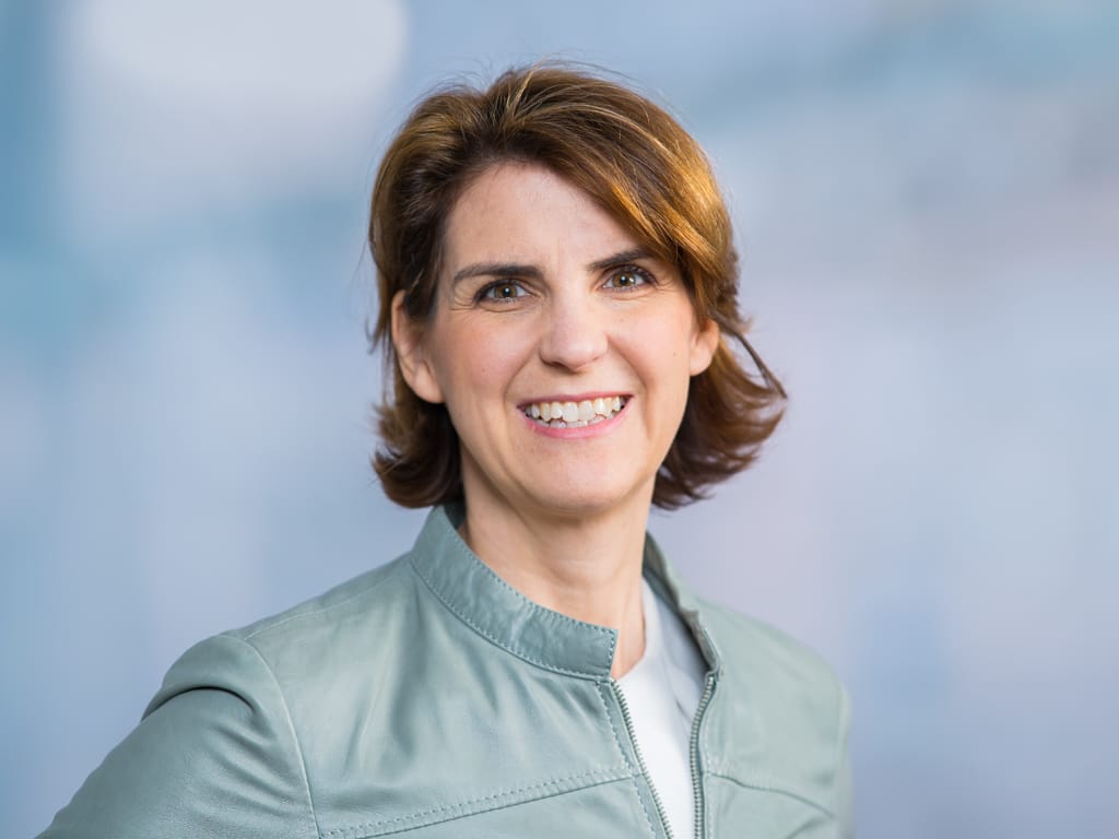 Person wearing a light green jacket over a white top with short brown hair against a blue background